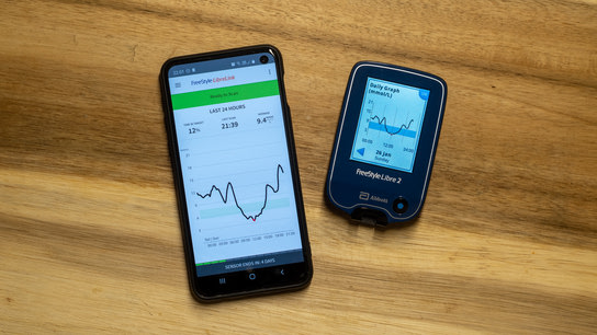 A smartphone with the LibreLink app next to the FreeStyle Libre Reader device. Both device show the same 24-hour glucose graph.