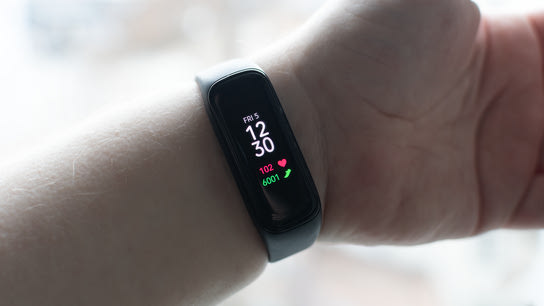 An outstretched arm showing off a Samsung Galaxy Fit2 activity armband. The armband is small with a bright display showing the current time, heart rate, and step count.
